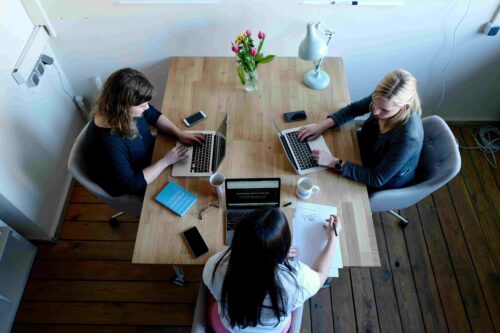 Three women collaborate at a table with laptops, coffee, and notes, brainstorming UGC social media strategies.